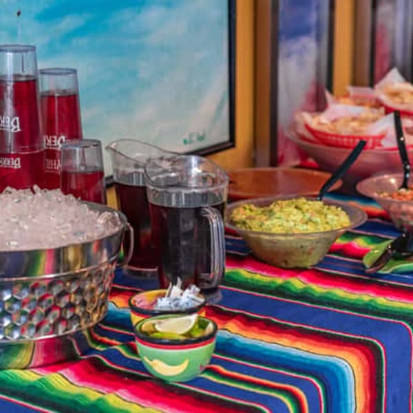 Mexican buffet with guacamole, salsa, chips, drinks, and a bucket of ice on a colorful striped tablecloth.